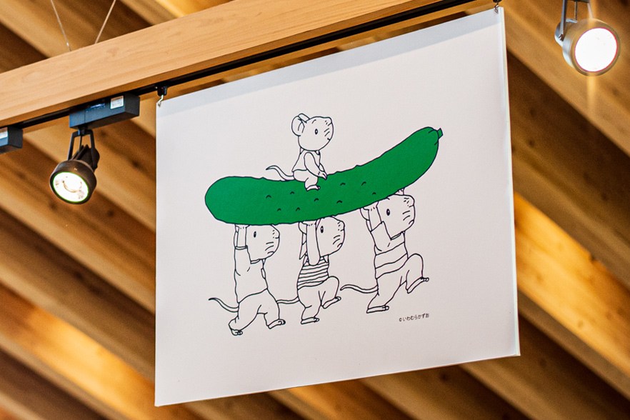 Illustration of cartoon characters carrying a large green cucumber, hanging from wooden ceiling with track lighting.