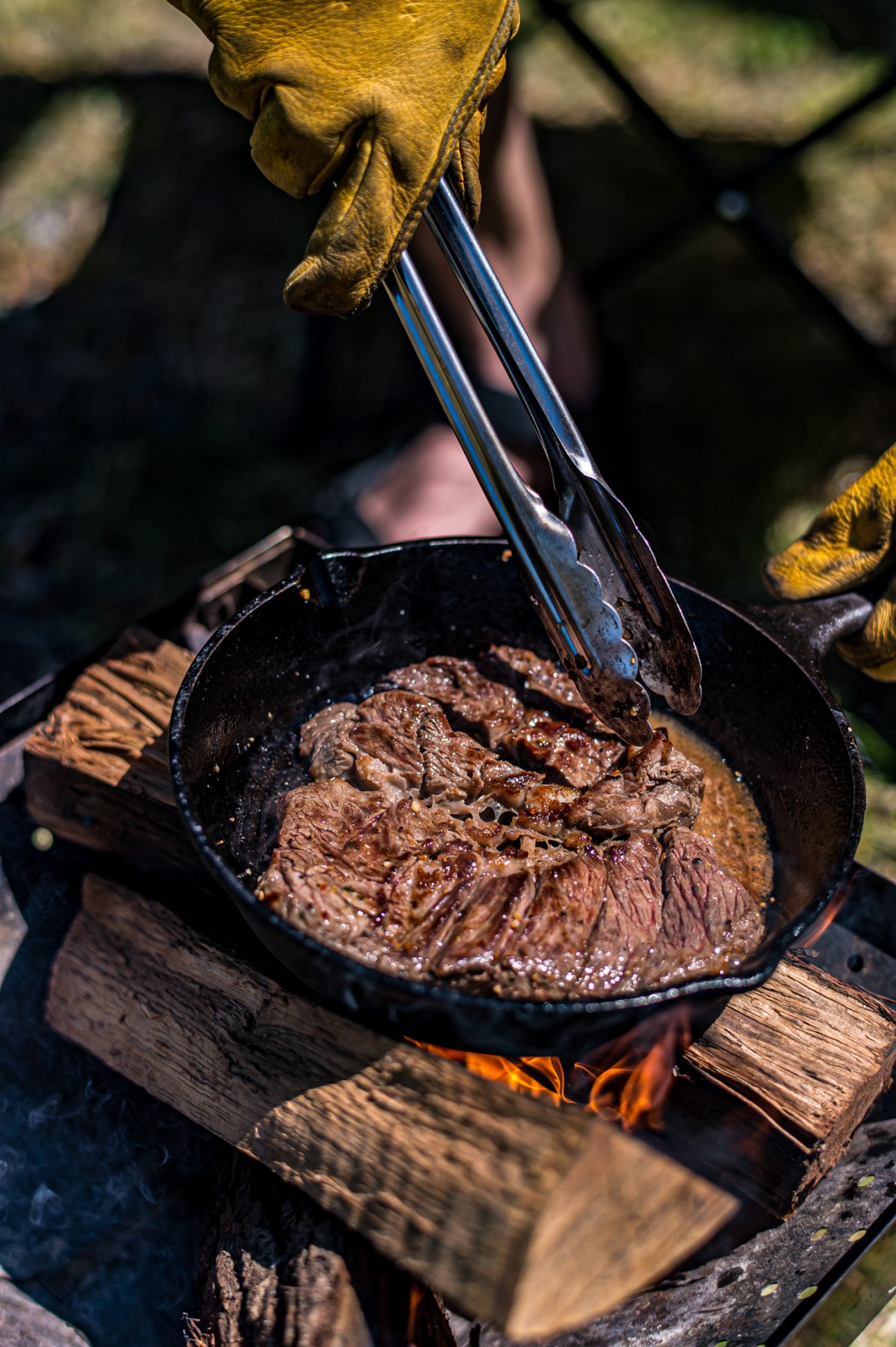 Hand in yellow leather gloves using tongs to handle cooked steak in cast iron pan over campfire