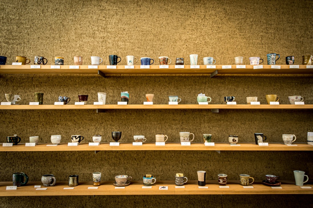 Wooden shelves displaying a collection of handmade ceramic cups and mugs by Mashiko potters against a cork wall.