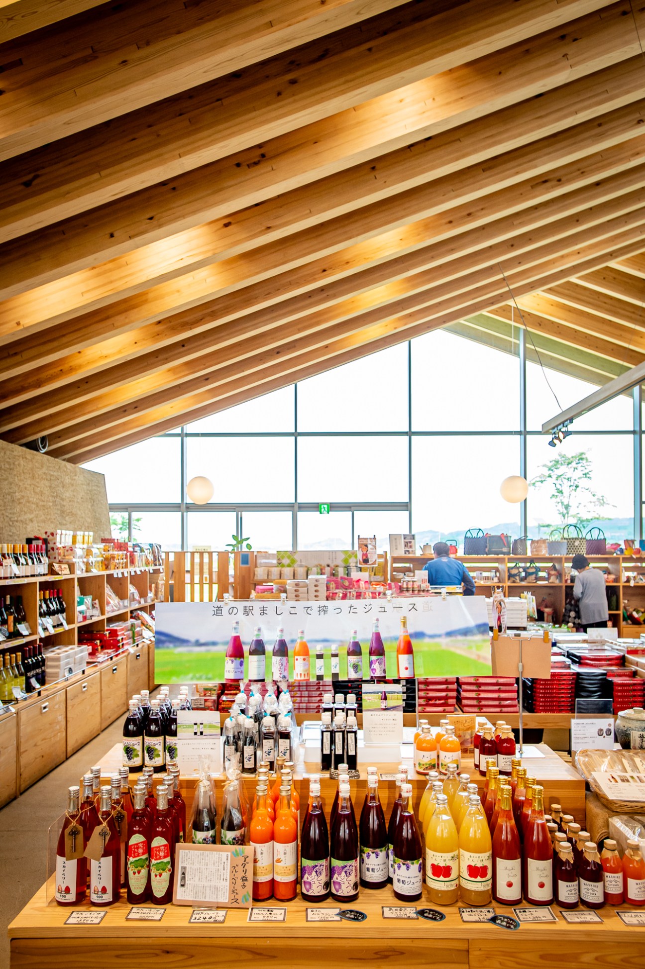 Interior of Mashiko Roadside Station displaying bottles of regional fruit juice on wooden counter with scenic rice field views through windows.