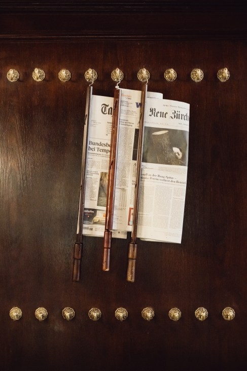 German newspapers hang from wooden holders on a dark wood wall decorated with brass ornamental knobs.