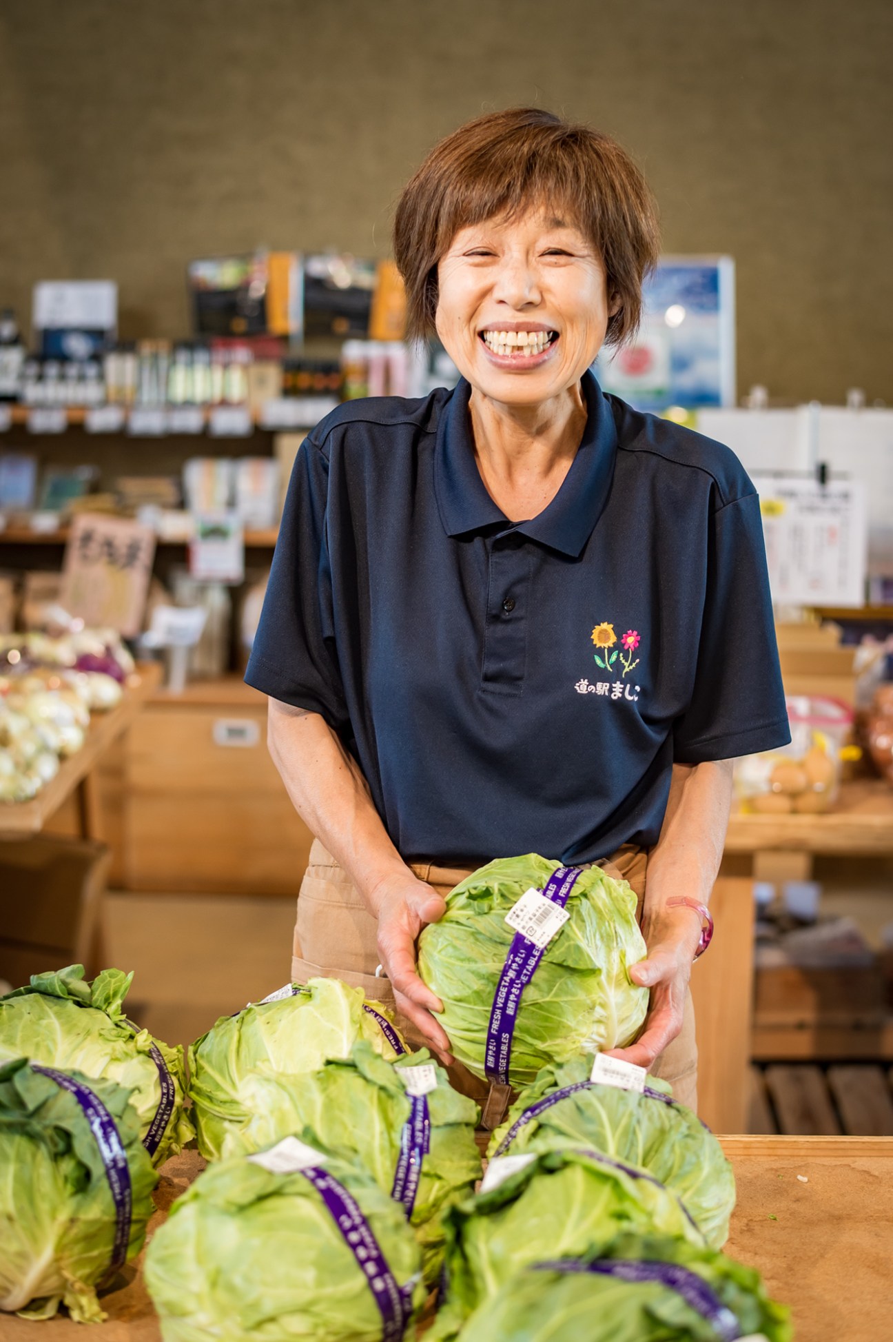 Smiling woman in navy polo shirt holding fresh cabbage at Mashiko Roadside Station farm shop.