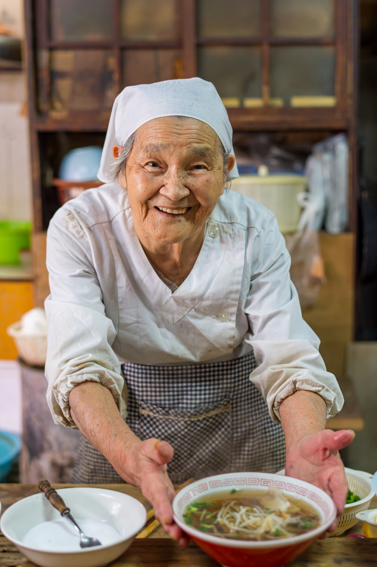 Elderly Japanese woman in white chef's coat and hat smiling while presenting a bowl of ramen soup