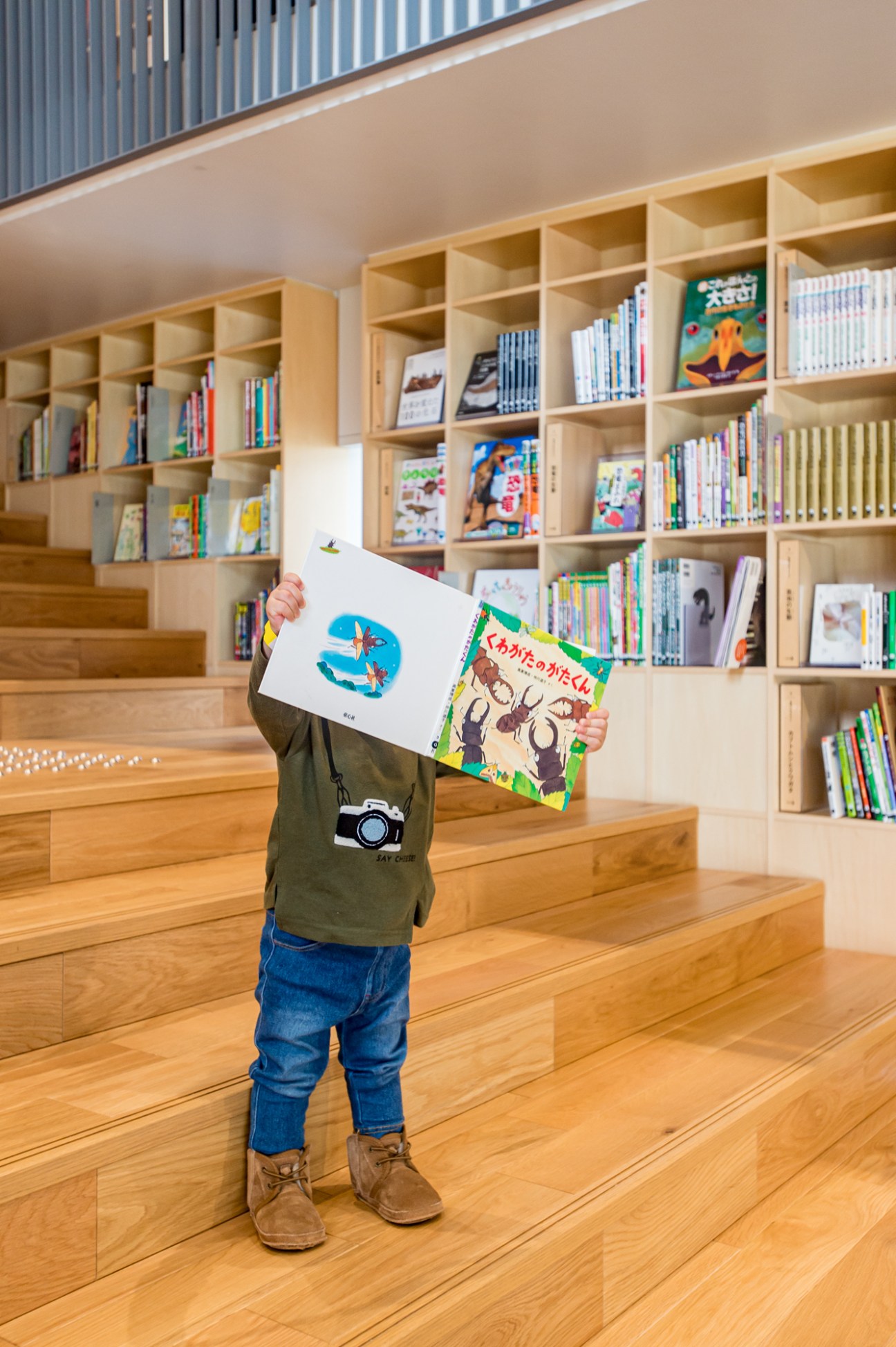 Child holding open picture books while standing on wooden steps in front of floor-to-ceiling bookshelves