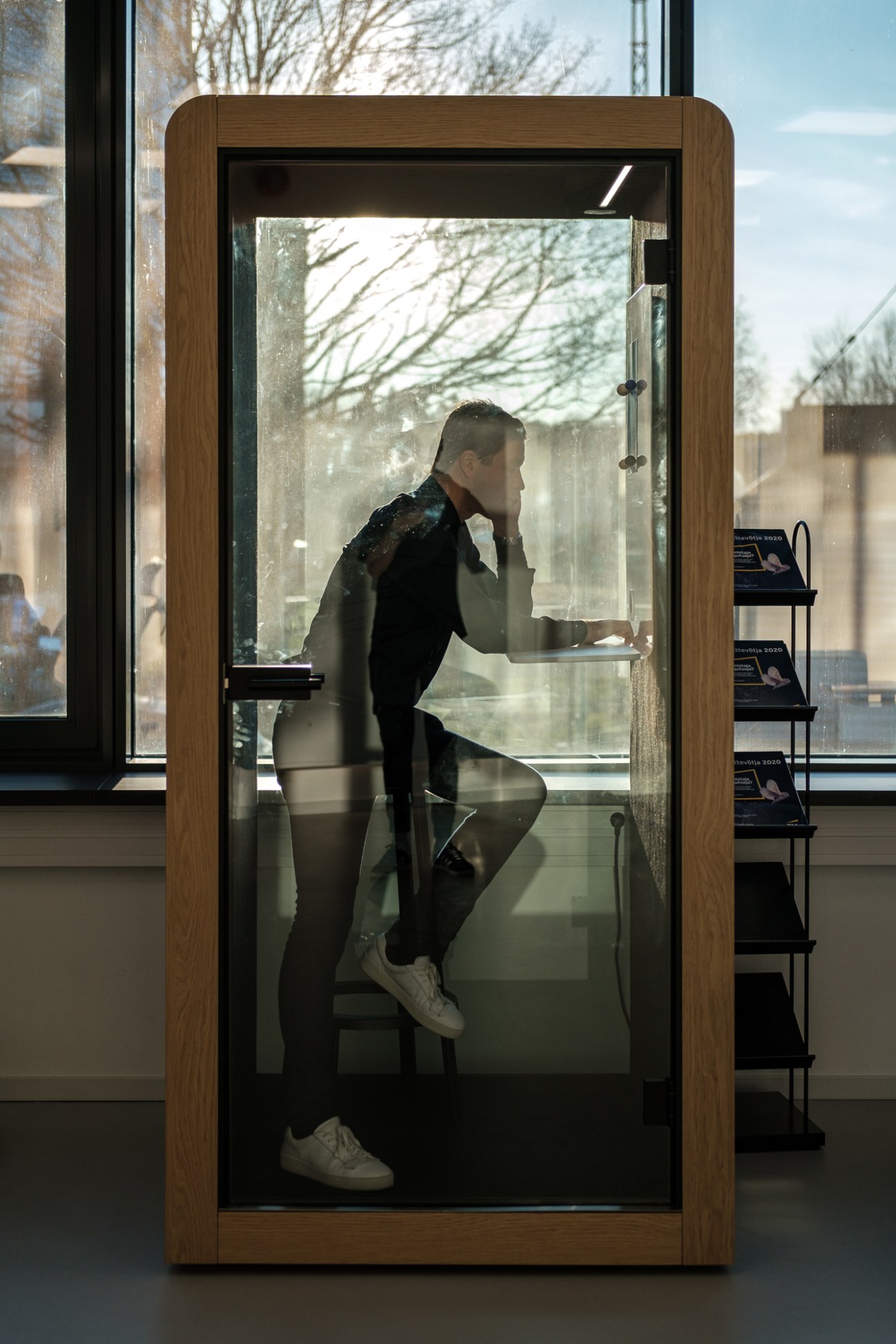 Person sitting inside a glass-walled Silen office pod with wooden frame, talking on phone in bright office space