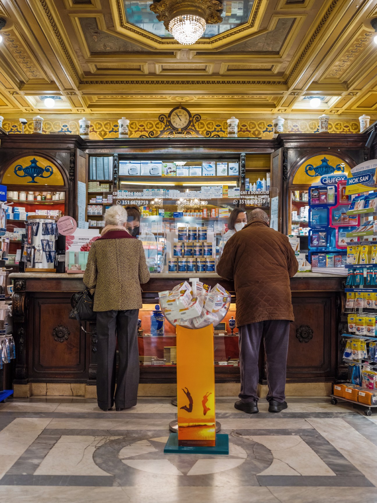 Interior of a historic Italian pharmacy with ornate wooden counter and shelving displaying medicinal products.