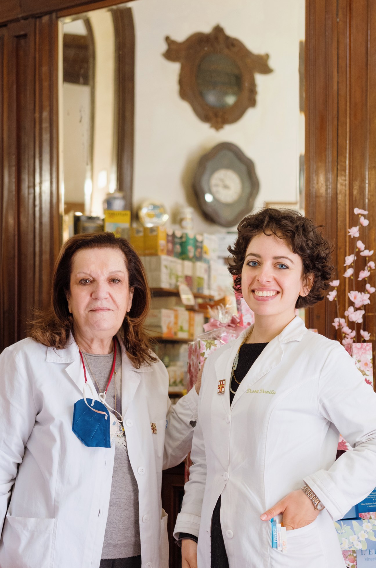 Two female pharmacists in white coats stand together in a pharmacy interior with vintage clocks on the wall behind them.
