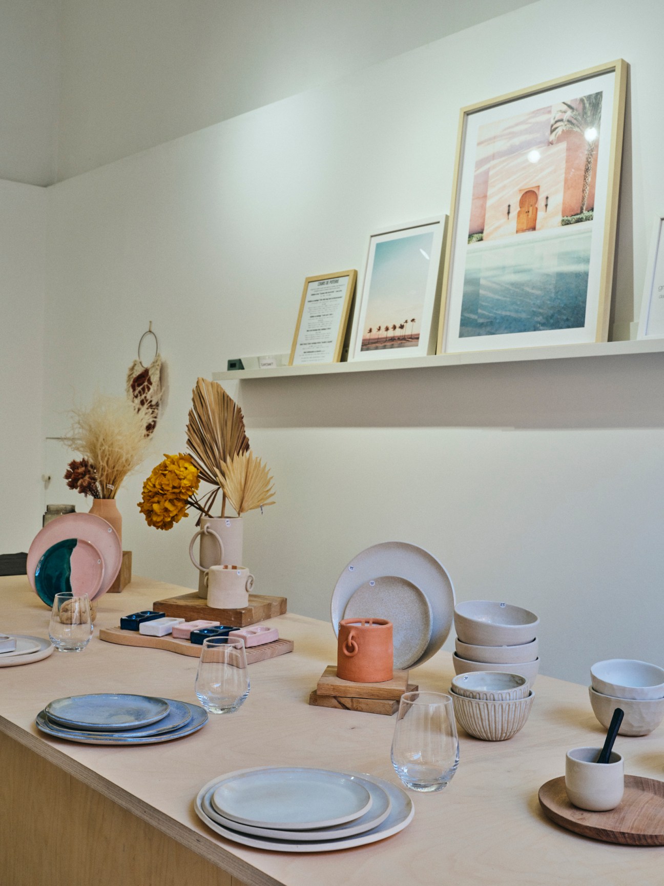 Artisanal ceramic tableware and dried flowers displayed on wooden counter with framed artwork on shelf above
