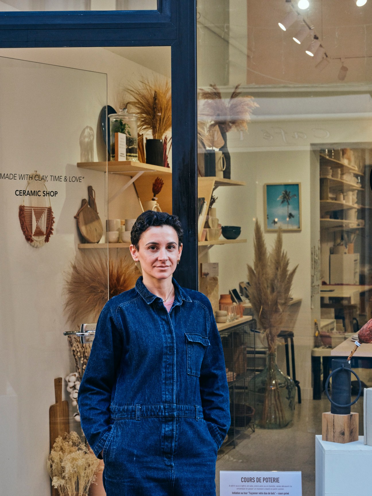 Woman in denim jumpsuit standing in front of ceramic shop window displaying pottery and crafts