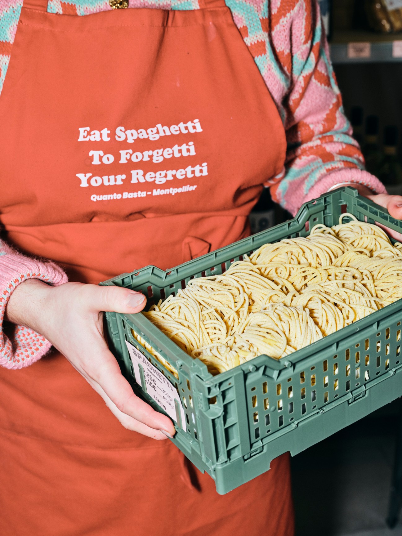 Person holding crate of fresh handmade pasta with humorous Italian apron reading 'Eat Spaghetti To Forgetti Your Regretti'