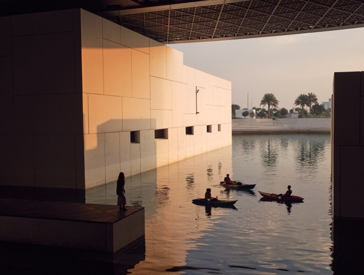 Jean Nouvel's geometric domed roof structure at the Louvre Abu Dhabi casting shadows over shallow water with boats.