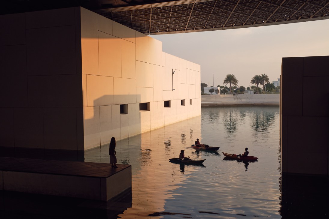 Jean Nouvel's geometric domed roof structure at the Louvre Abu Dhabi casting shadows over shallow water with boats.