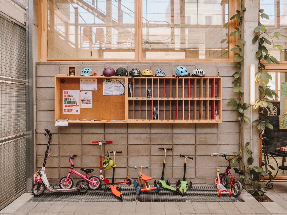 A wooden noticeboard and storage wall with cork bulletin board, shelving cubbies, bicycle helmets, children's bikes, scooters and climbing equipment in a communal courtyard area.