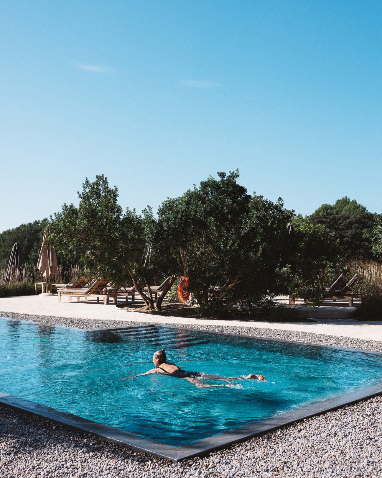 Person swimming in blue pool surrounded by wooden loungers, umbrellas and lush Mediterranean trees