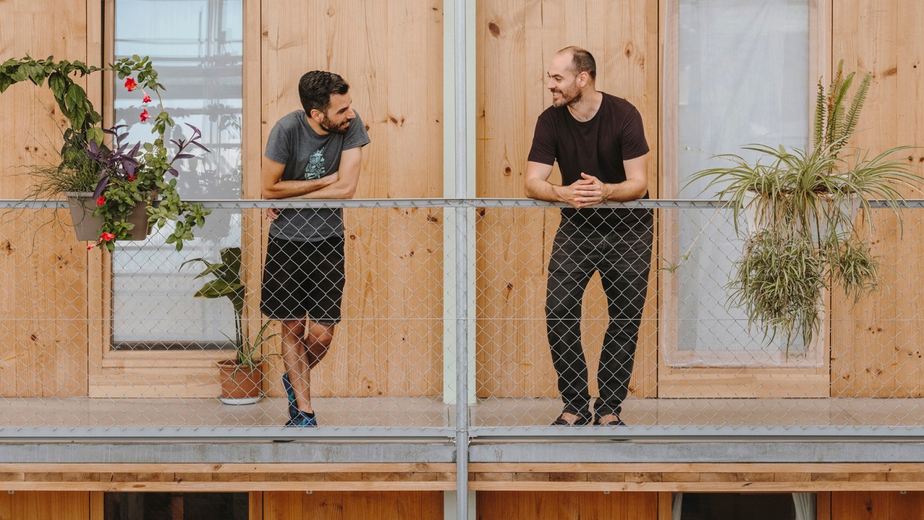 Two men stand on a wooden balcony with potted plants, smiling at each other in conversation.