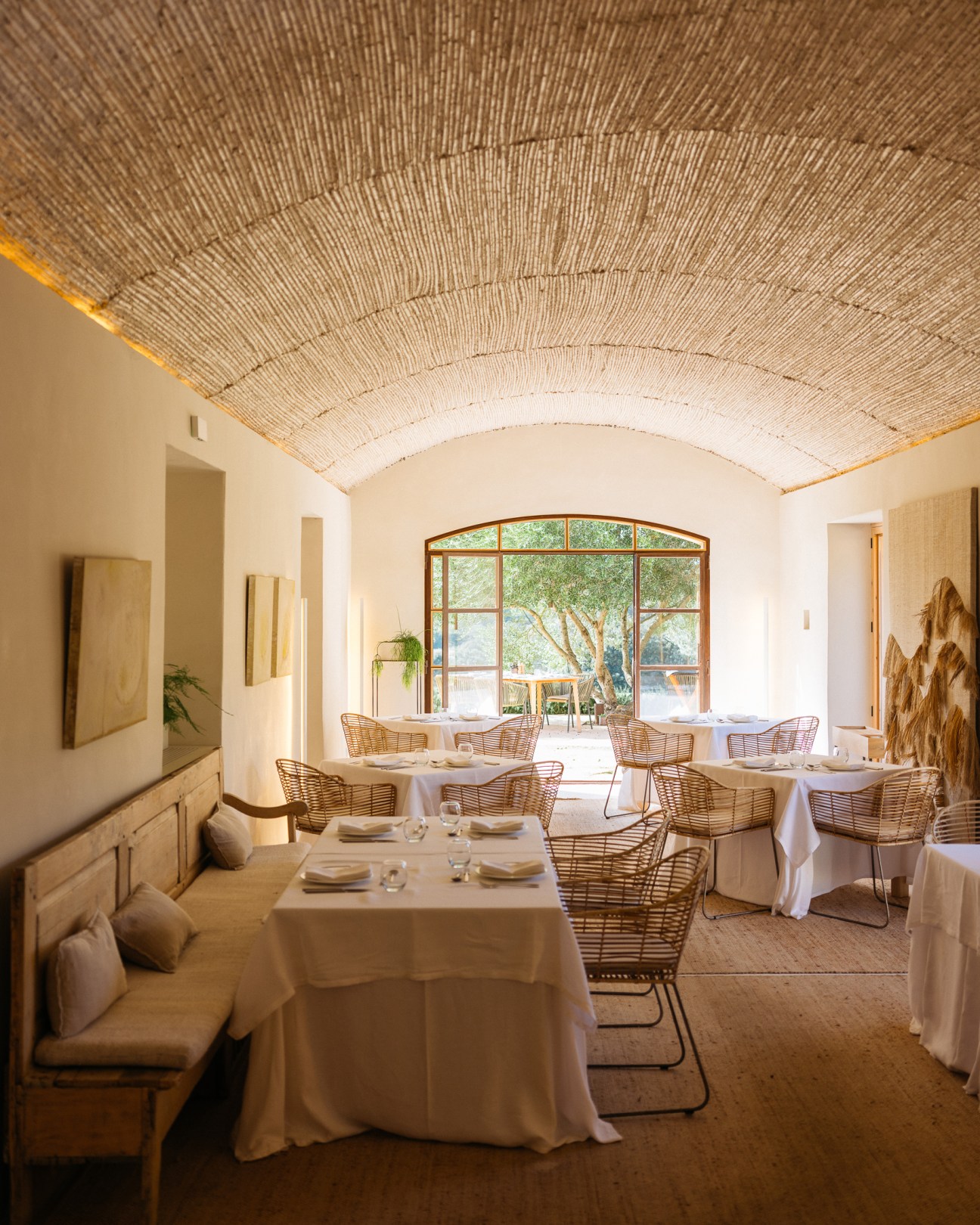 Restaurant dining room with exposed brick vaulted ceiling, wicker chairs, white tablecloths and arched window