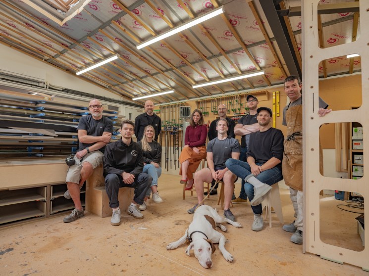Team photo of Young & Norgate furniture makers and collaborators gathered in their Bristol workshop with a dog