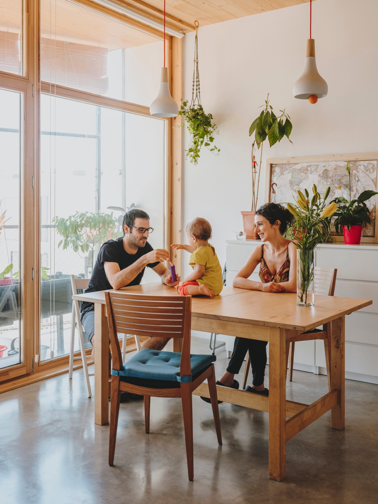 Family of three seated at wooden table in bright apartment with potted plants and large windows overlooking balcony.