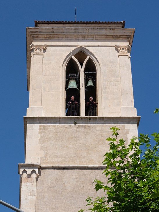 Stone bell tower with Gothic arched windows housing two bronze bells, with two people visible inside the belfry at San Silvestro belltower