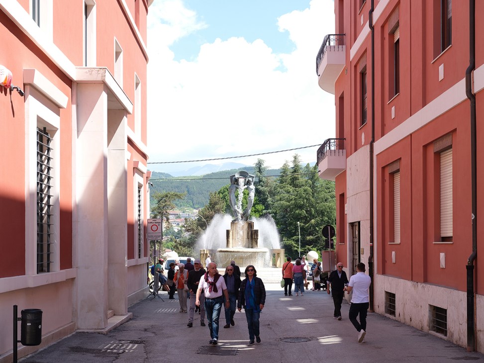 People walking down a street between coral-colored buildings toward a fountain Fontana Luminosa and the Apennine mountains in background