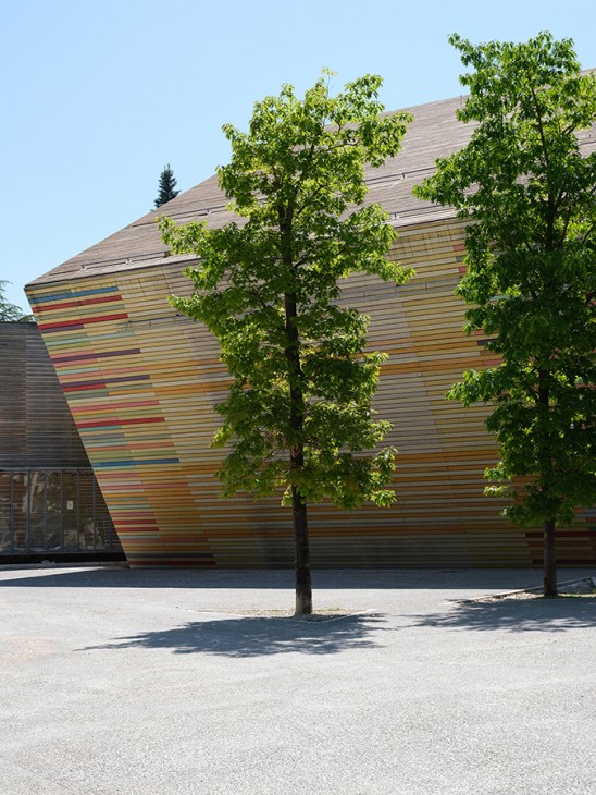 Modern curved building with colorful horizontal striped facade behind leafy green trees on sunny street at Renzo Piano