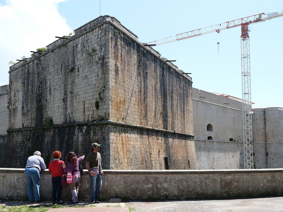 Tourists observe ancient stone fortress walls at Forte Spagnolo with construction crane visible in background during restoration work