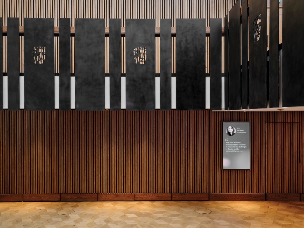Modern interior wall with wood slat ceiling, dark vertical panels featuring mask-shaped motifs, and digital display