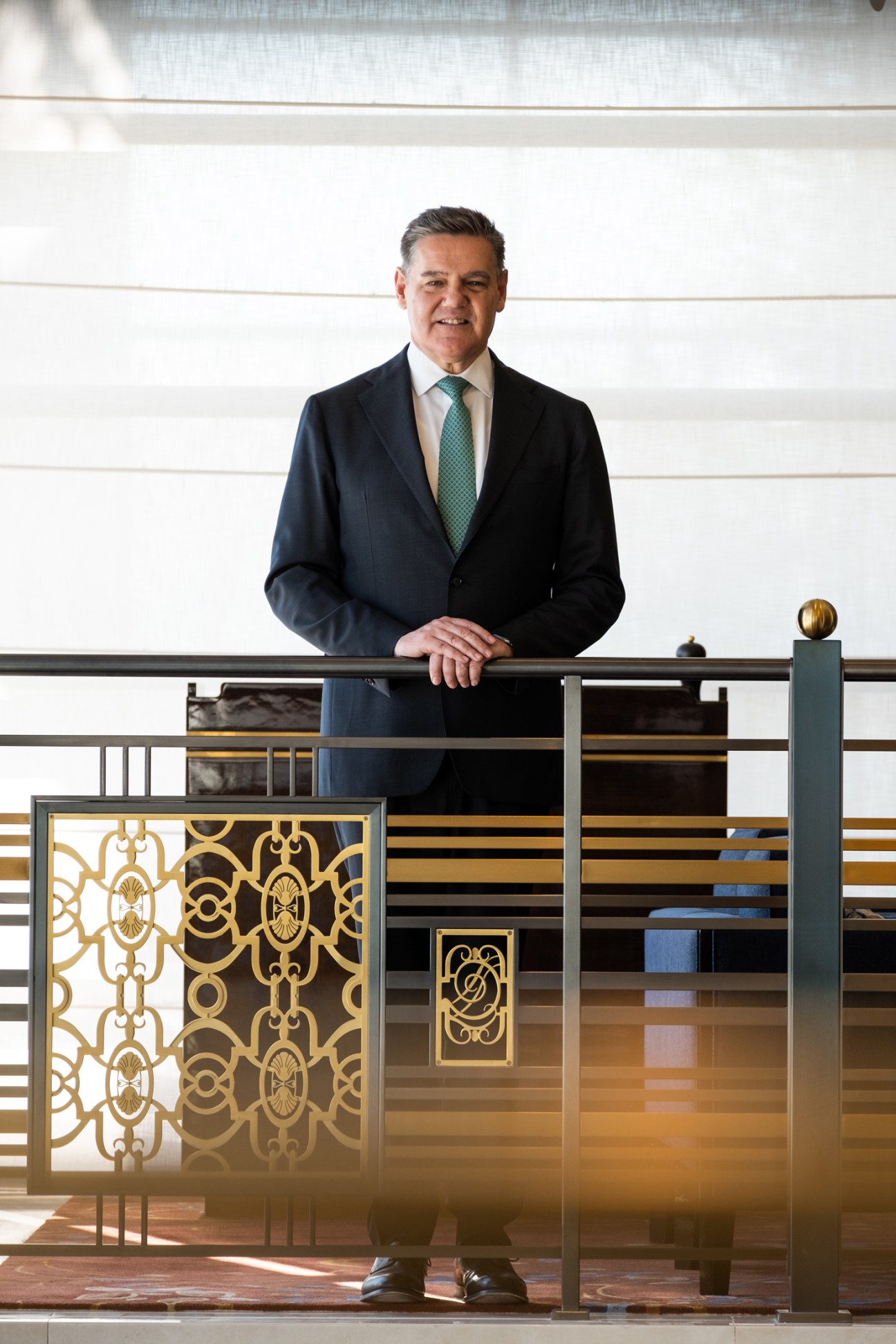 Man in dark suit and green tie standing at ornate hotel balcony railing with decorative gold metalwork