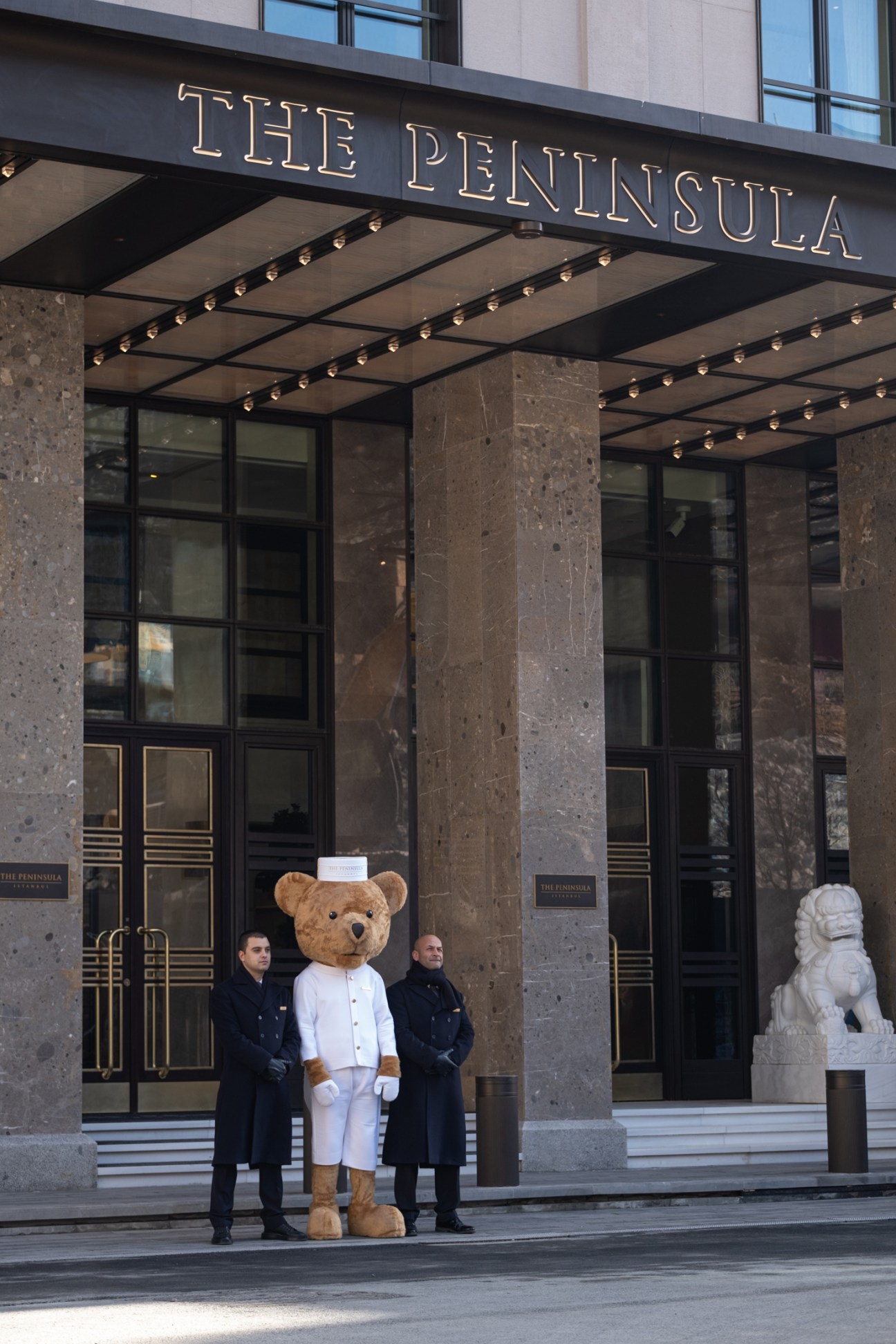 Large teddy bear mascot in white chef's uniform and hat standing with hotel staff at The Peninsula hotel entrance