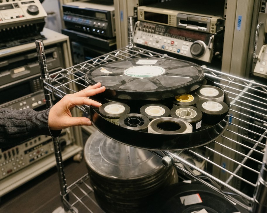 A hand holds a circular film reel with subtitles stamped onto it in a dubbing studio with professional audio equipment in the background.