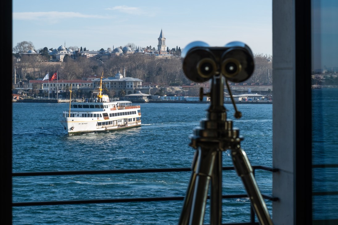 View through hotel window showing binoculars on tripod overlooking Bosphorus with ferry boat and Istanbul skyline