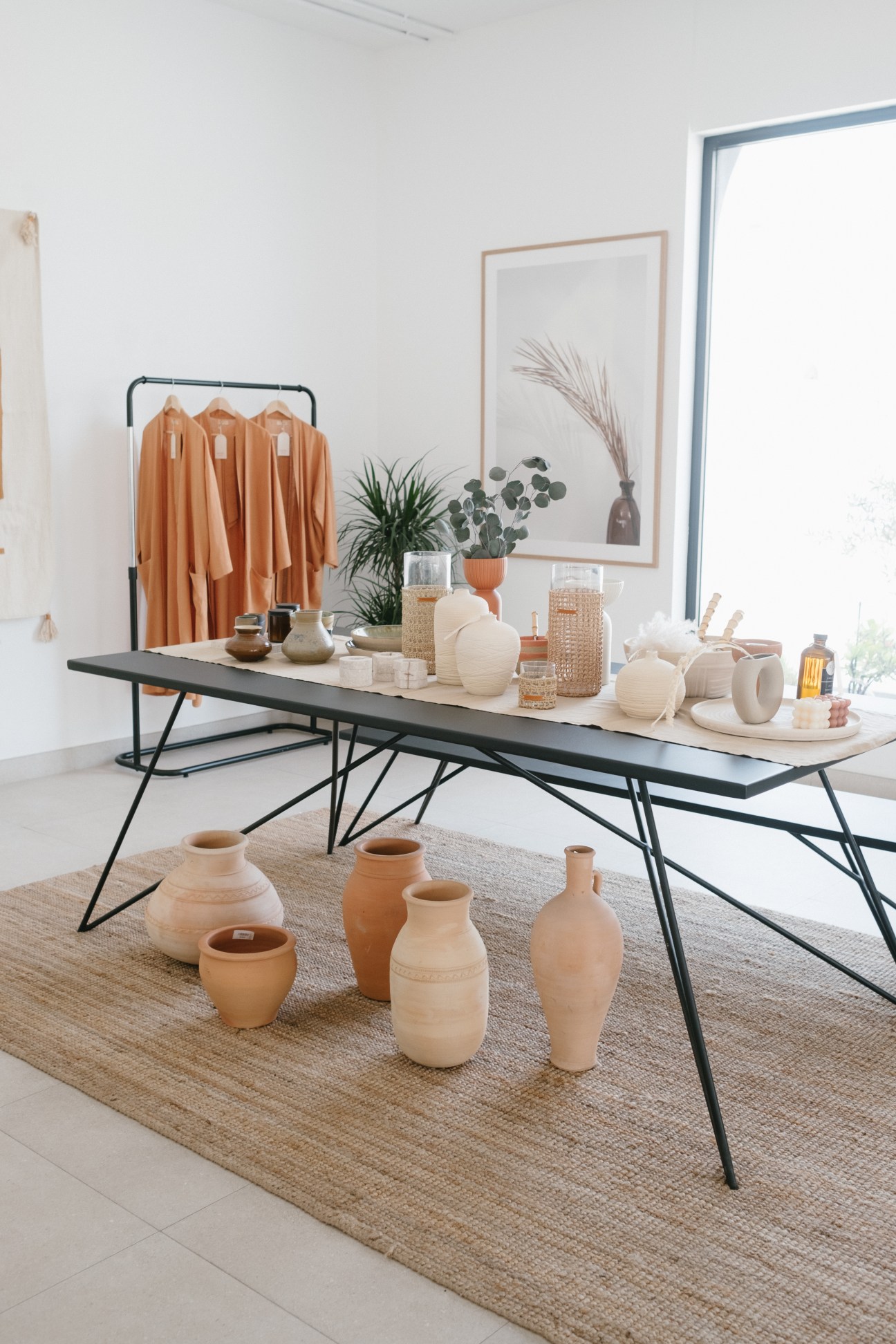 Modern table display with terra cotta pottery, clothing rack, plants and home goods in minimalist interior