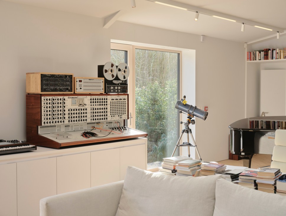Modern studio workspace with vintage synthesizer, reel-to-reel tape machine, telescope, and forest views through large windows.