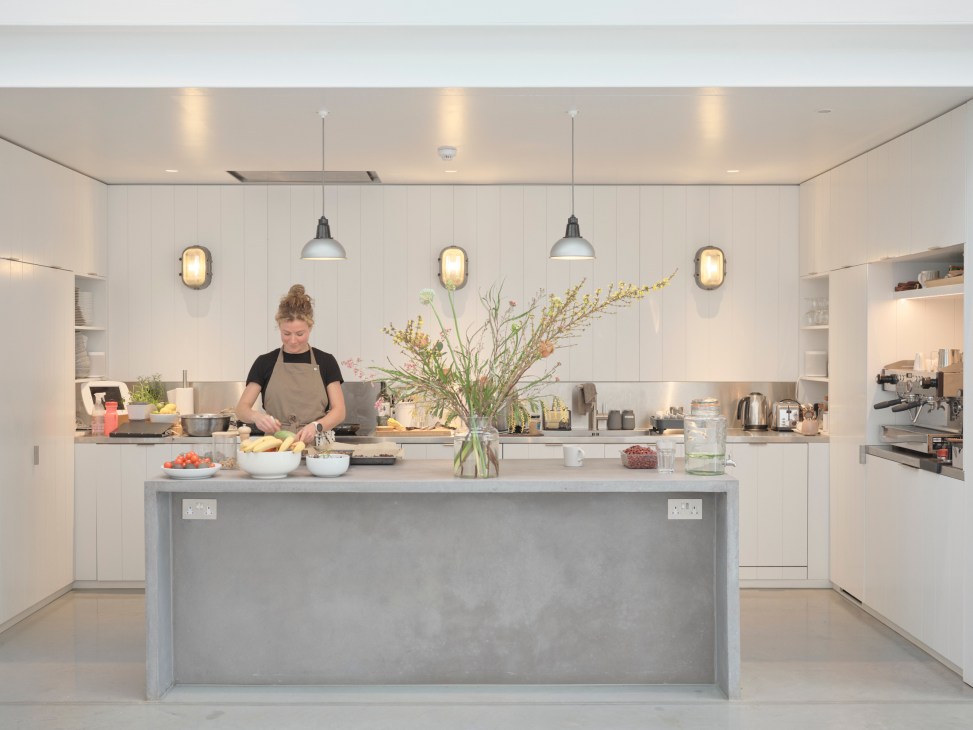 Woman in apron preparing food in a modern kitchen with white cabinetry, concrete island, and pendant lighting.