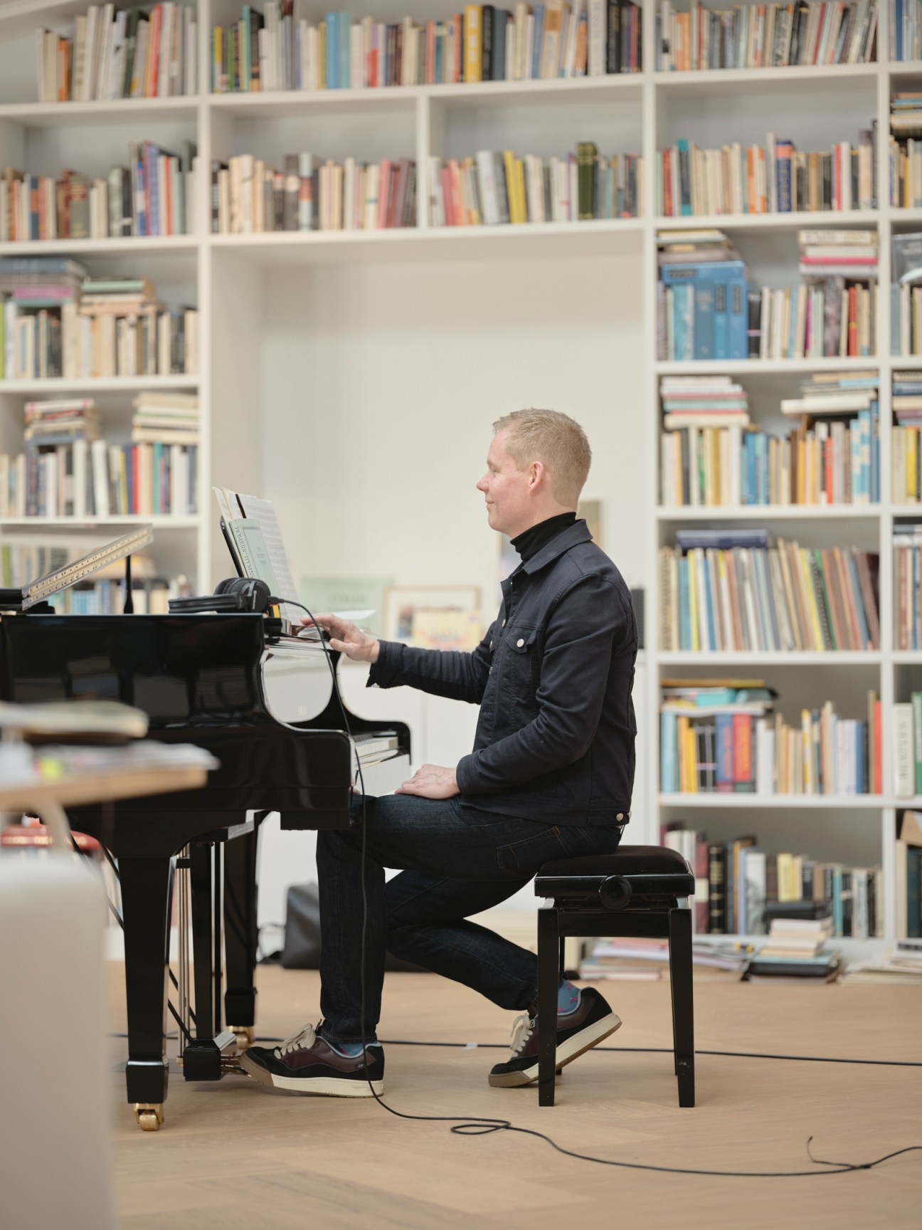 Composer Max Richter sits at a black grand piano in a bright studio space with white bookshelves behind him.