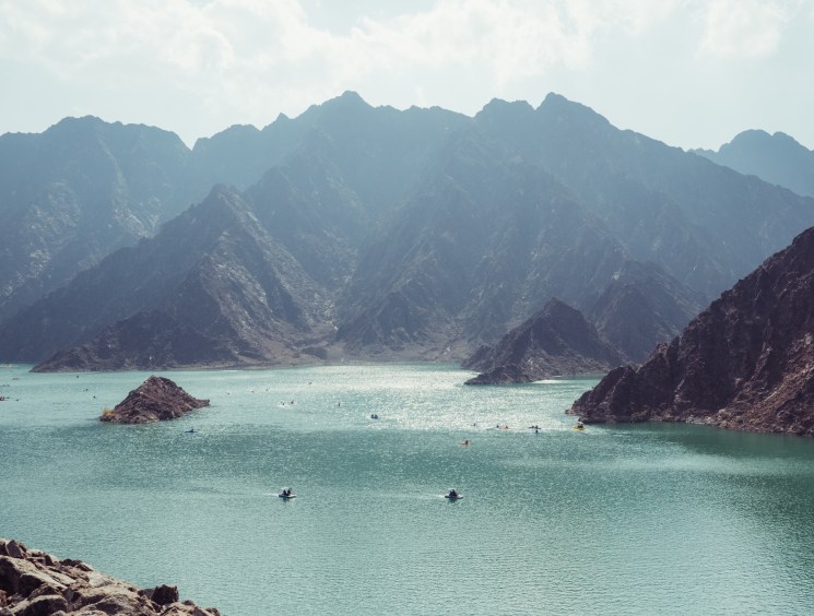 Turquoise waters of Hatta Dam surrounded by rugged Hajar mountain peaks with swimmers and kayakers visible.