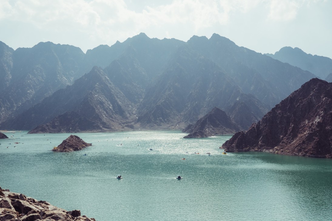 Turquoise waters of Hatta Dam surrounded by rugged Hajar mountain peaks with swimmers and kayakers visible.