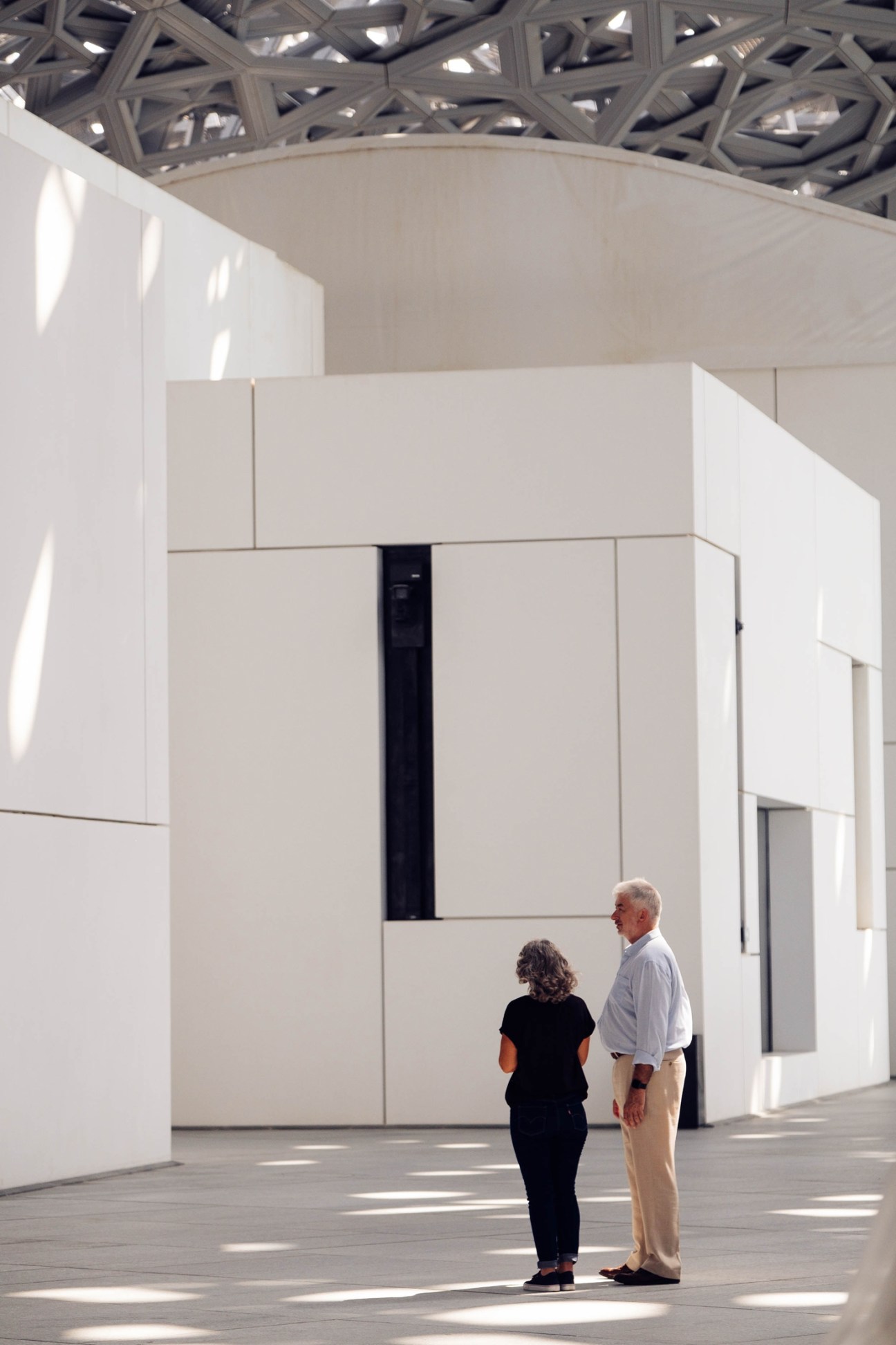 Two visitors stand in a bright gallery space beneath an ornate geometric ceiling at the Louvre Abu Dhabi.