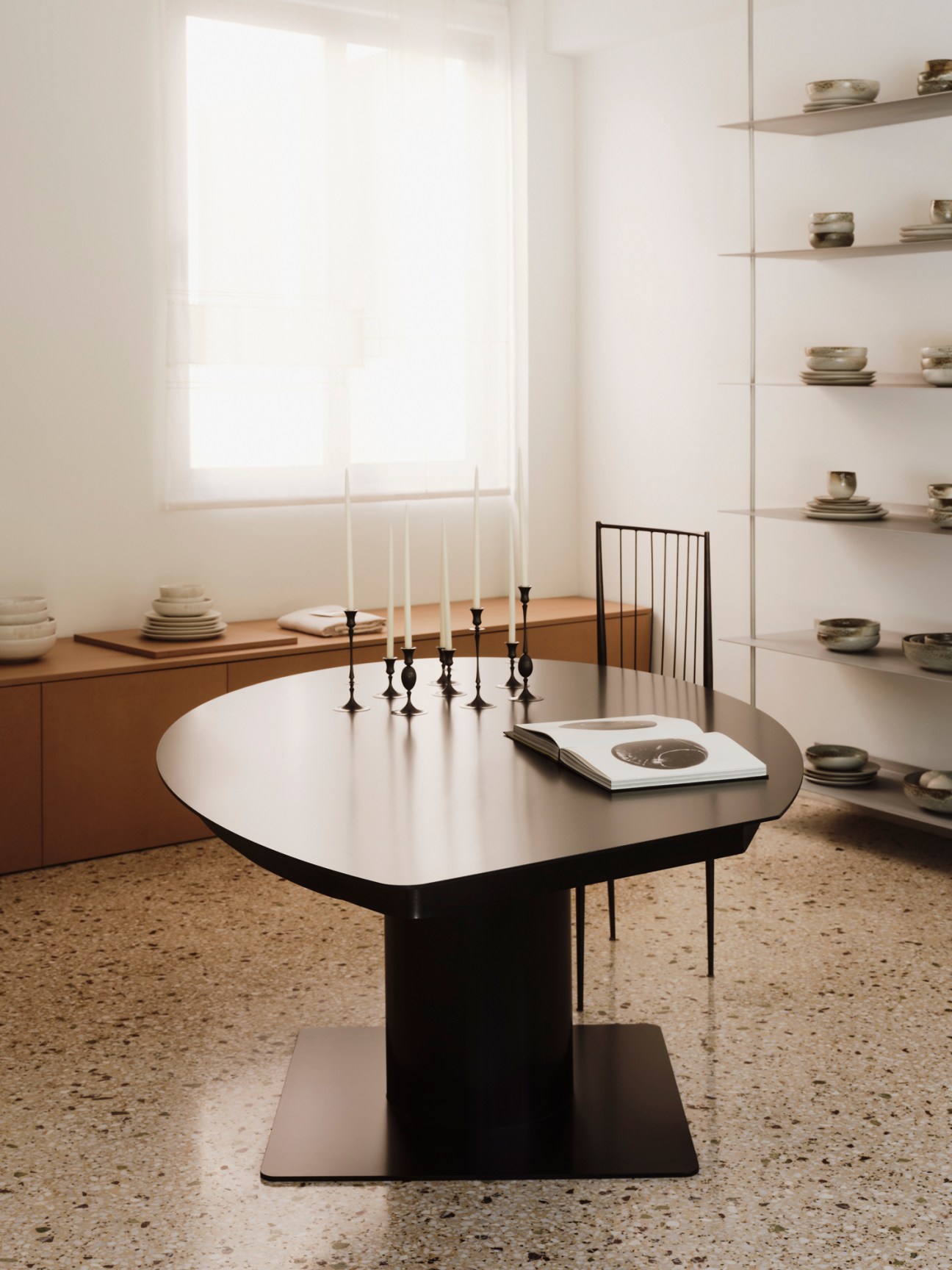 Dark oval dining table with candlesticks and book on top, single chair visible in minimalist room with shelving