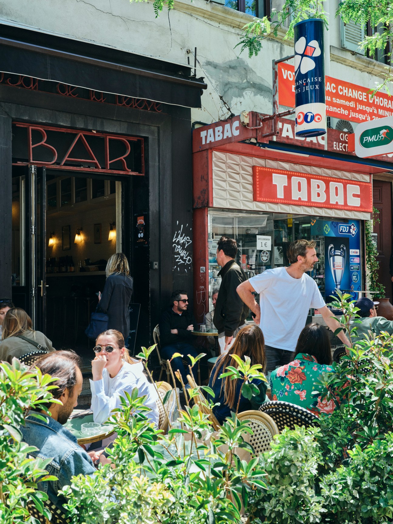 Outdoor terrace seating with diners in front of a bar and tobacco shop (Tabac) on a Marseille street, with potted plants and ivy visible