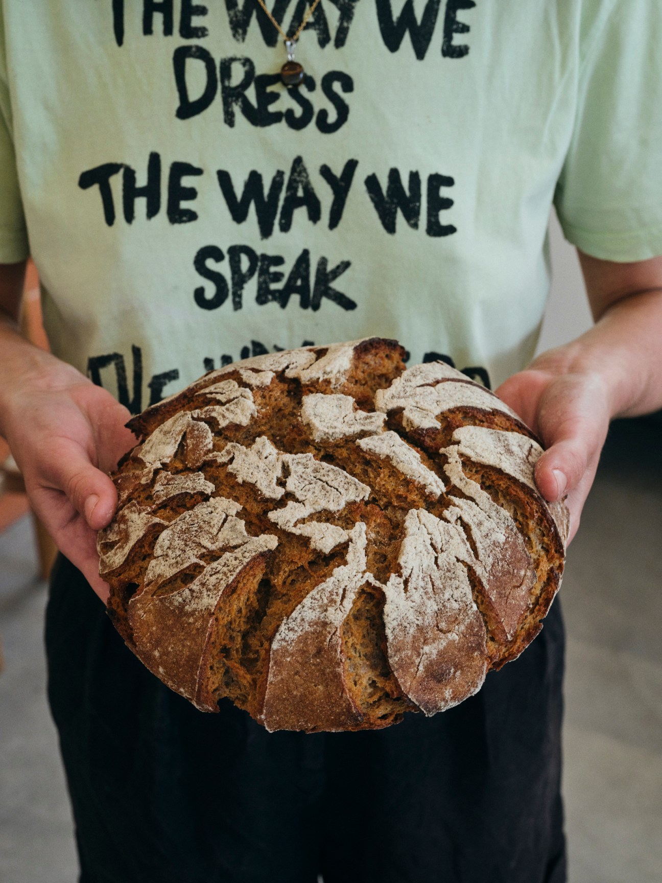Person in white t-shirt holding a rustic artisanal sourdough bread loaf with decorative scoring pattern