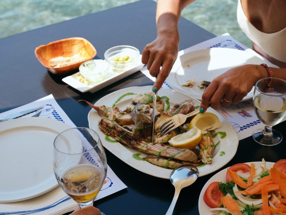 Person cutting grilled fish at seaside restaurant table with salad and wine