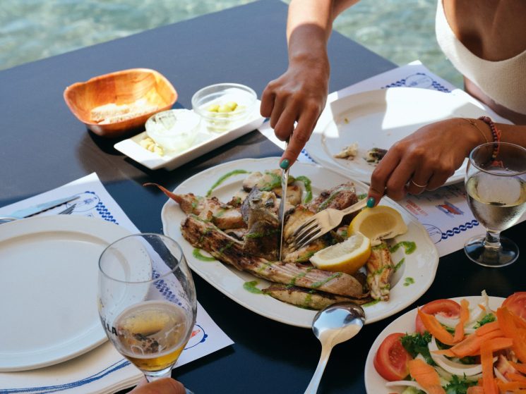 Person cutting grilled fish at seaside restaurant table with salad and wine