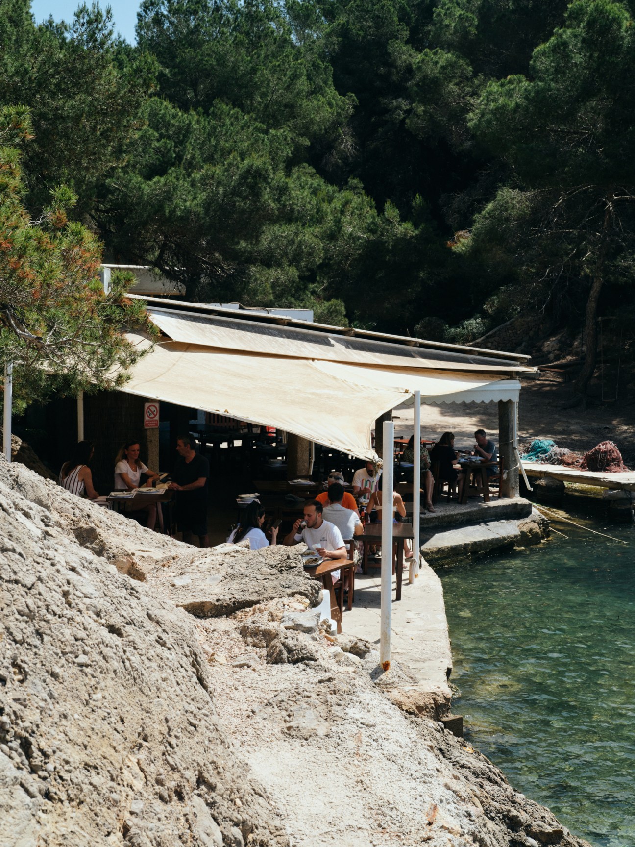 Rustic beachside restaurant with canvas awning, diners at tables, and boats moored in clear turquoise water
