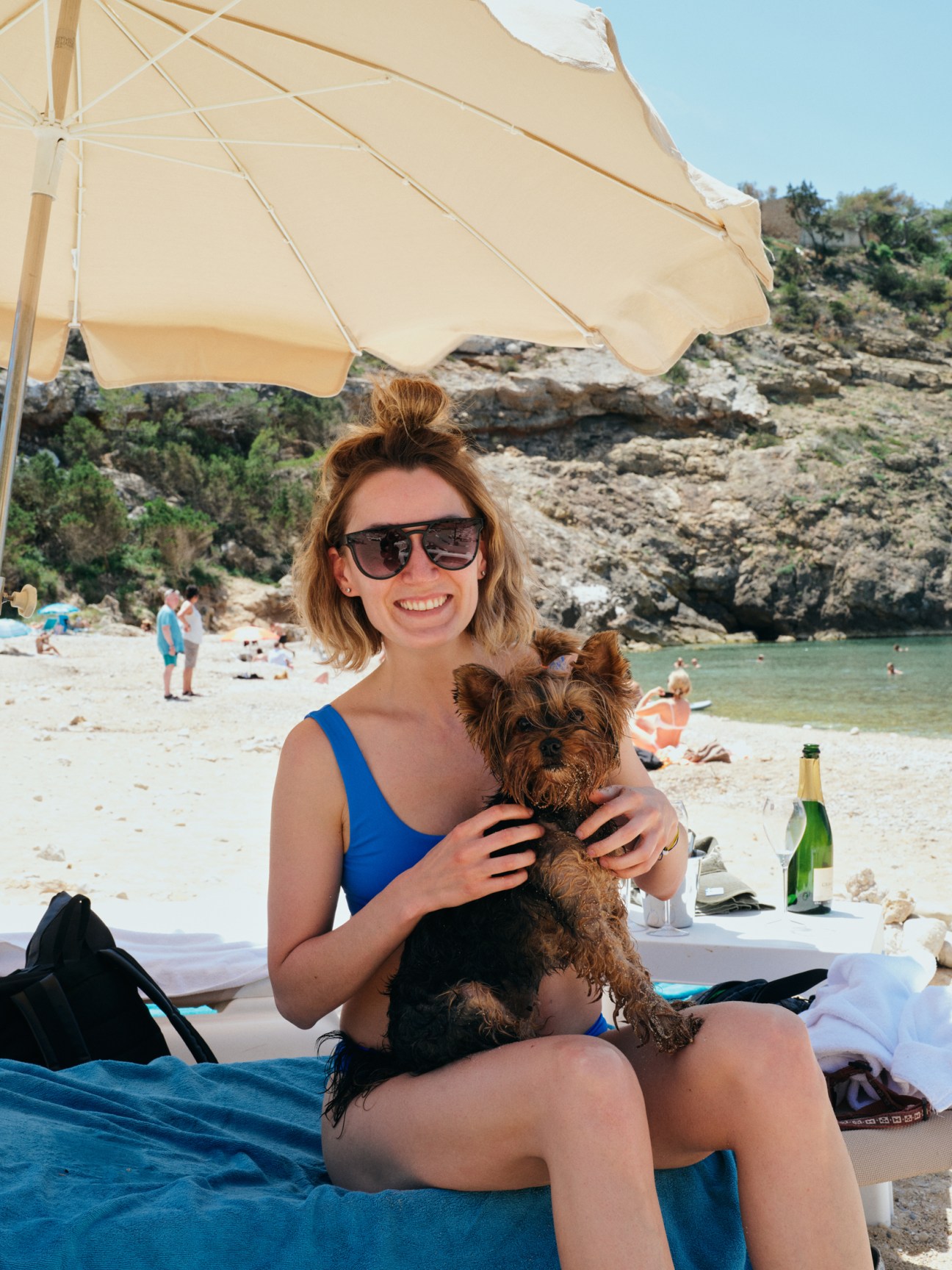 Woman in blue swimsuit holding small Yorkshire Terrier dog while sitting under beach umbrella