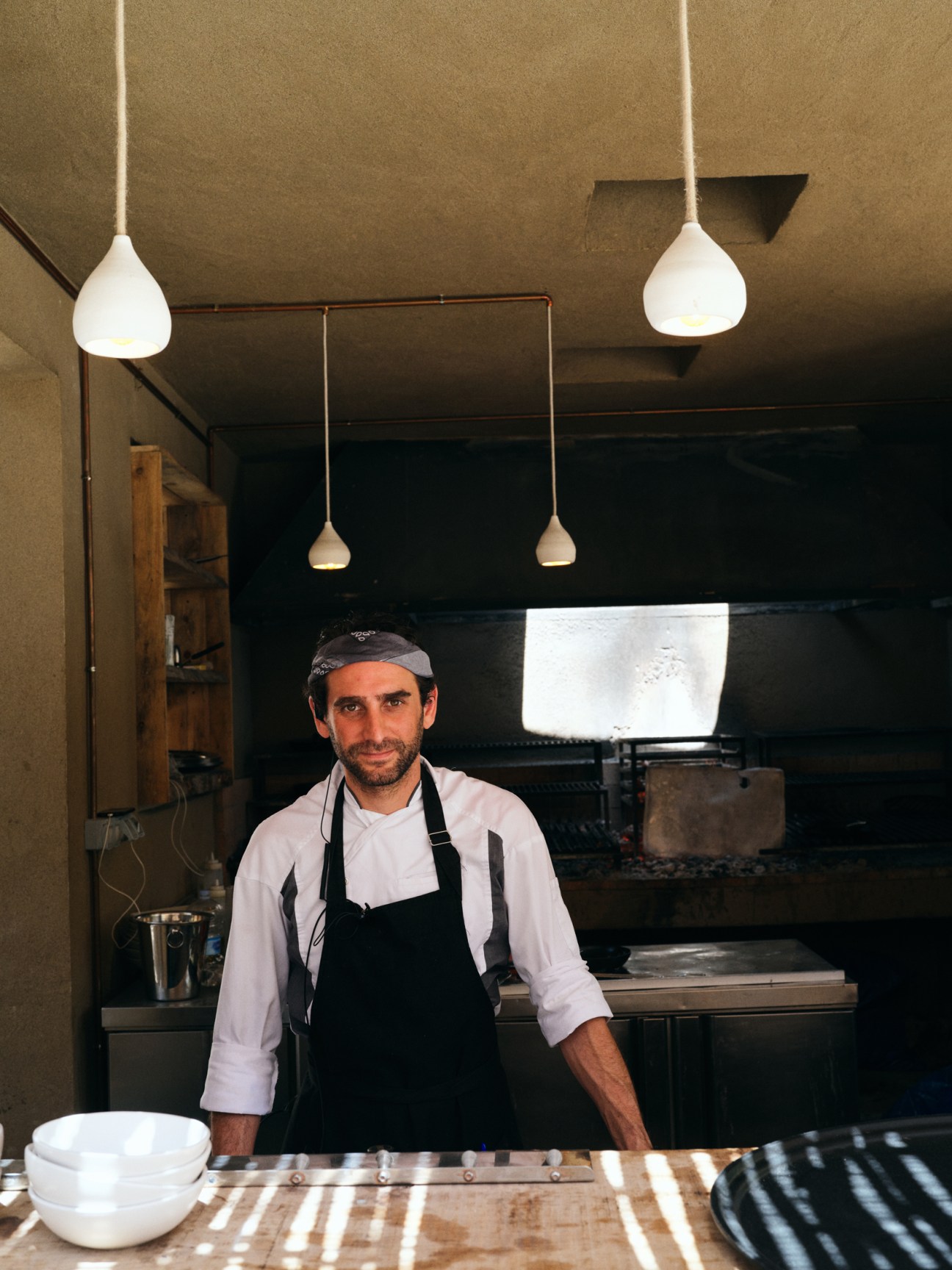 Chef in white jacket and black apron standing in restaurant kitchen with pendant lights overhead