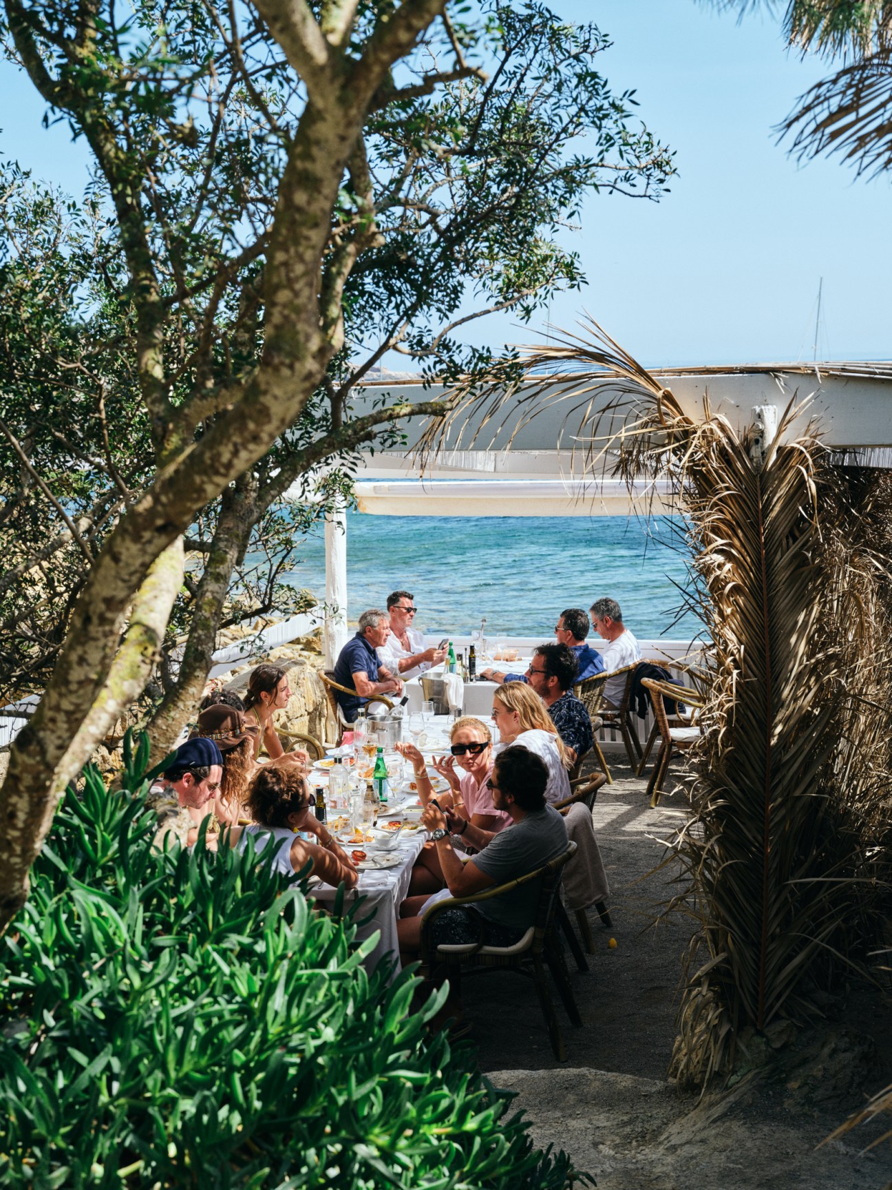 Diners enjoying lunch at beachside restaurant tables surrounded by tropical palm fronds overlooking blue water