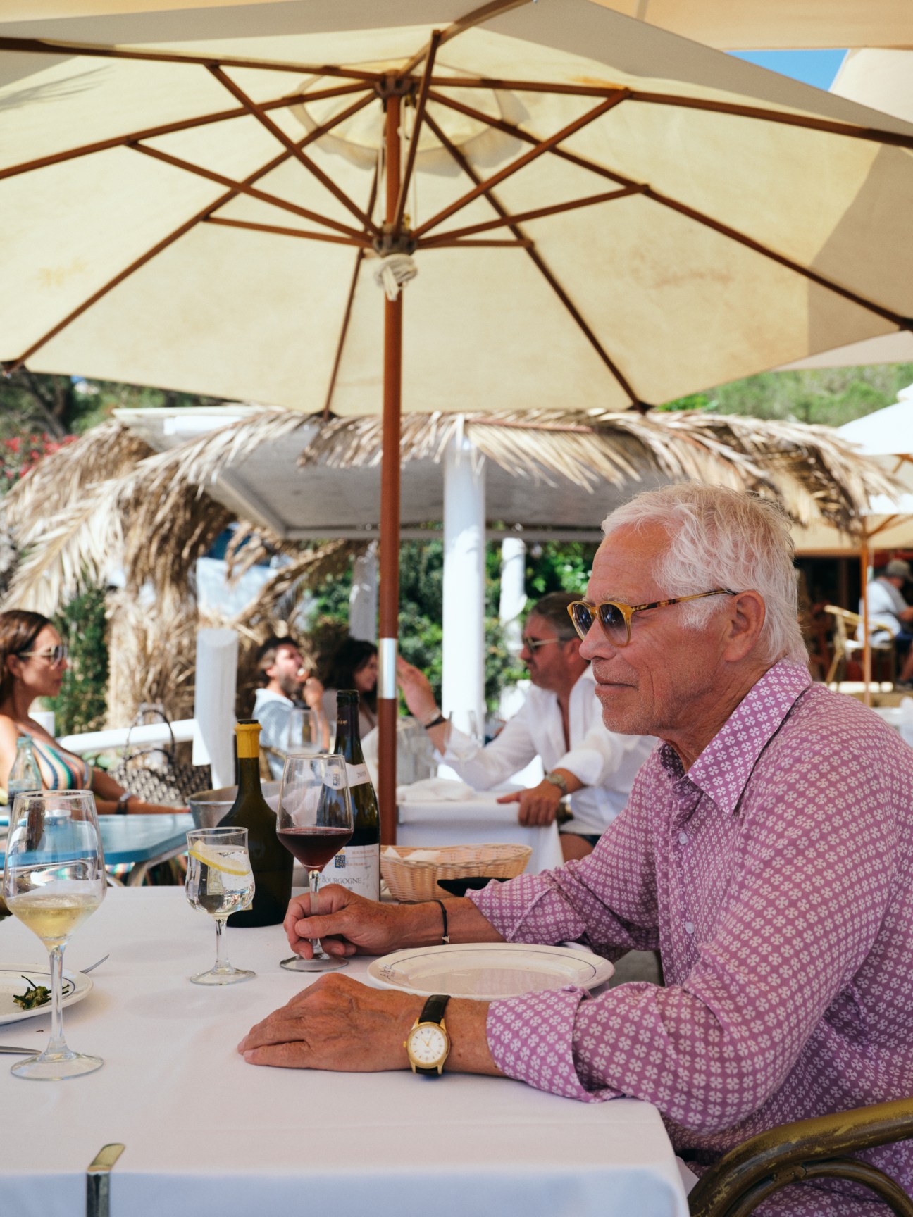 Elderly man with white hair and sunglasses sitting at outdoor restaurant table with wine glass under umbrella