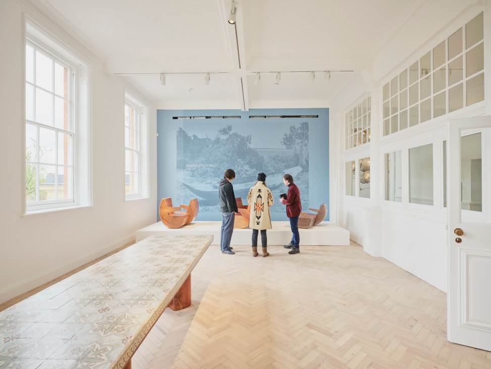 Three visitors viewing orange sculptural seats and a blue landscape artwork in a bright gallery space with white walls and large windows.