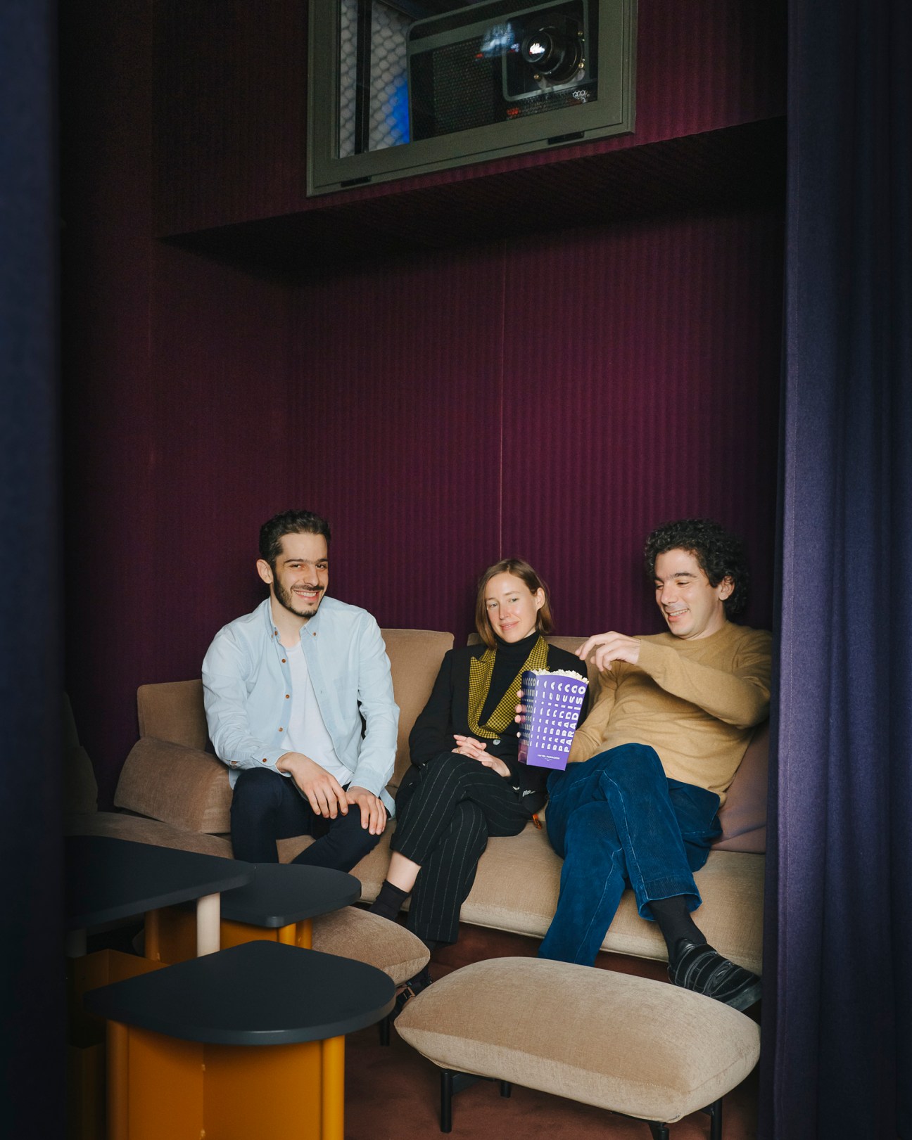 Three people sitting on a beige sofa in a cinema room with burgundy walls and a screen visible above.